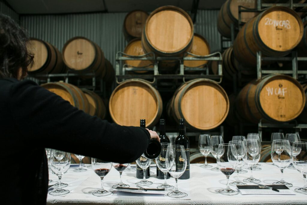 Back view of anonymous pommelier pouring cider from bottle into wineglass while standing at table with glassware in cidery against barrels